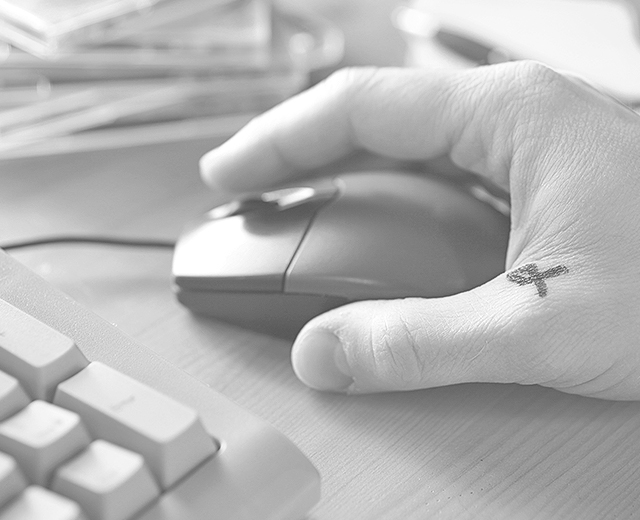 Ein Schreibtisch mit einer Tastatur und einer Maus. Auf der Maus ist eine Hand mit einem kleinen Tattoo zwischen Daumen und Zeigefinger.